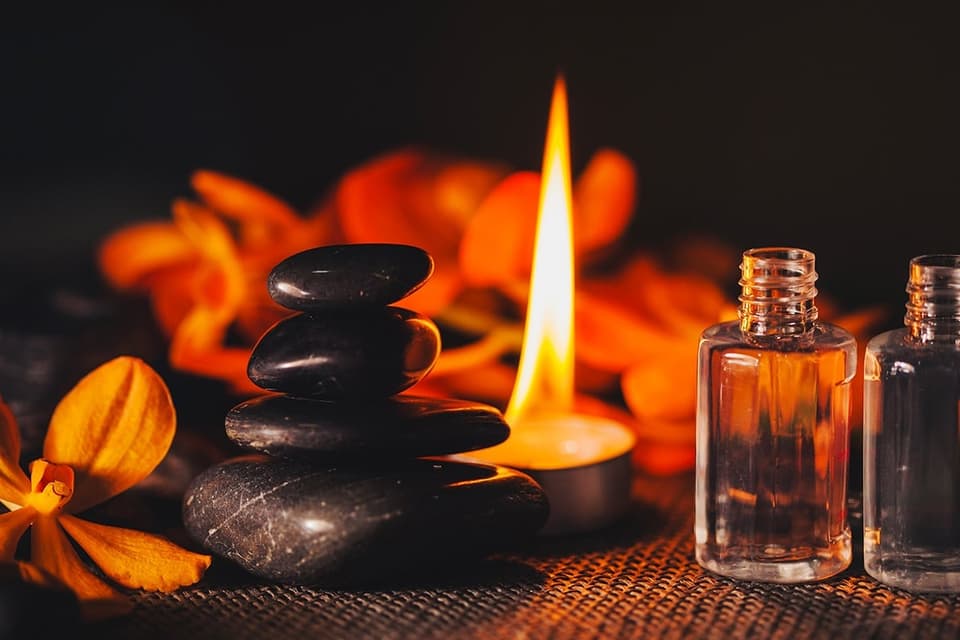 Gestapelte dunkle Massagesteine, brennende Kerze, Öl-Fläschchen und orange Blüten